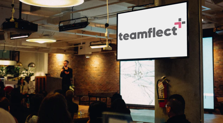 a man presenting something on stage with a teamflect logo on presentation screen